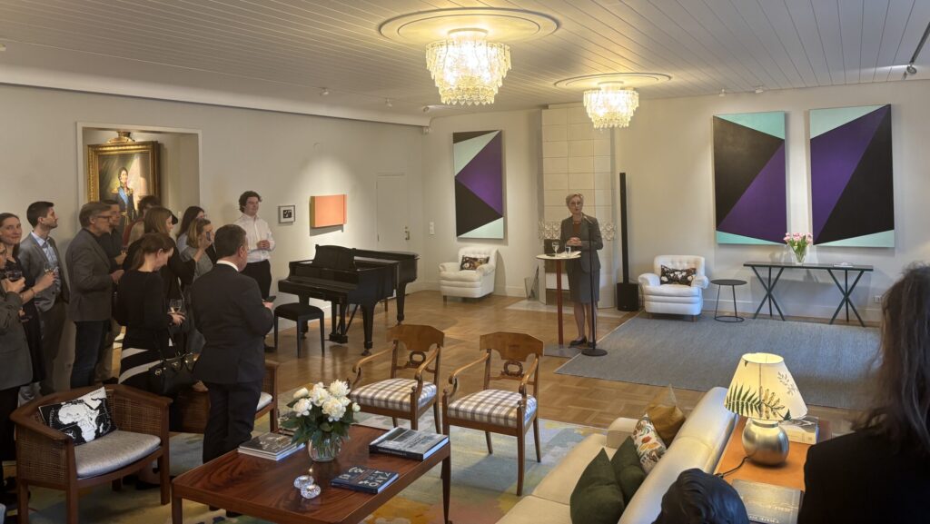 A group of people stands and listens to a speaker at a lectern in an elegant drawing room with chandeliers, a grand piano, modern art on the walls, and comfortable seating.