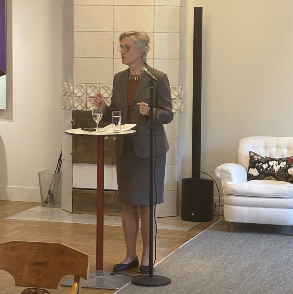 A woman in a grey suit speaks at a microphone stand in a well-lit room with a white sofa, a rug, and a tiled fireplace behind her. She stands at a small round table with a glass of water and holds a pen.