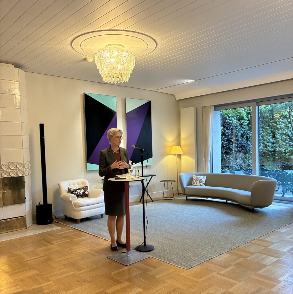 A woman in a suit stands at a microphone and lectern, speaking in a modern, well-lit room with wooden floors, contemporary furniture, and abstract art on the wall. Large windows reveal greenery outside.