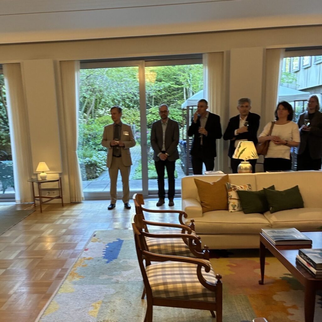 A group of people in formal attire stand near large windows in a spacious, well-lit lounge with patterned rugs, wooden chairs, and a white sofa. The windows reveal a green garden outside.