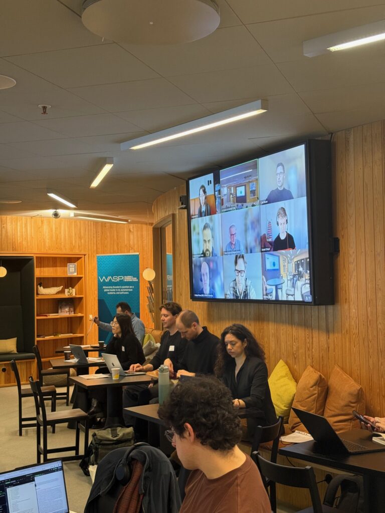 People are working on laptops in a modern meeting room with wood-panelled walls. A large screen shows a video call with multiple participants. Some people are seated, others are focused on their computers.