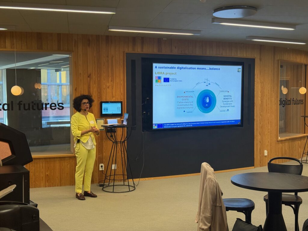 A woman in a yellow suit stands and presents in front of a screen displaying a slide titled “A sustainable digitalisation means… balance” in a modern room with wood-panelled walls and “digital futures” written on the glass.