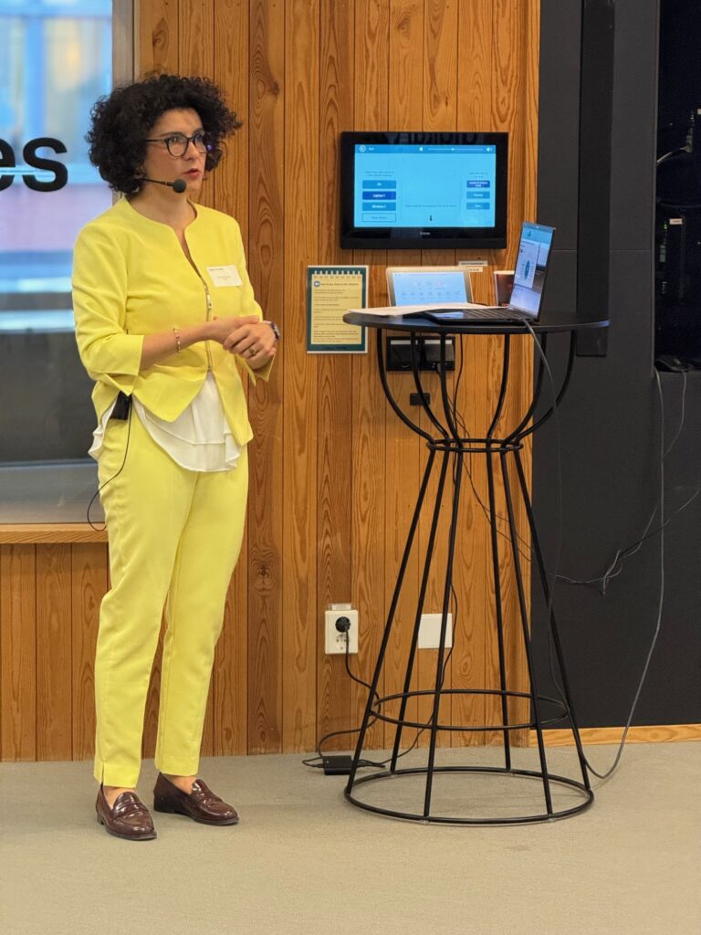 A woman in a yellow suit stands next to a high round table with a laptop, giving a presentation. She wears glasses and a headset, and a screen with charts is mounted on the wooden wall behind her.