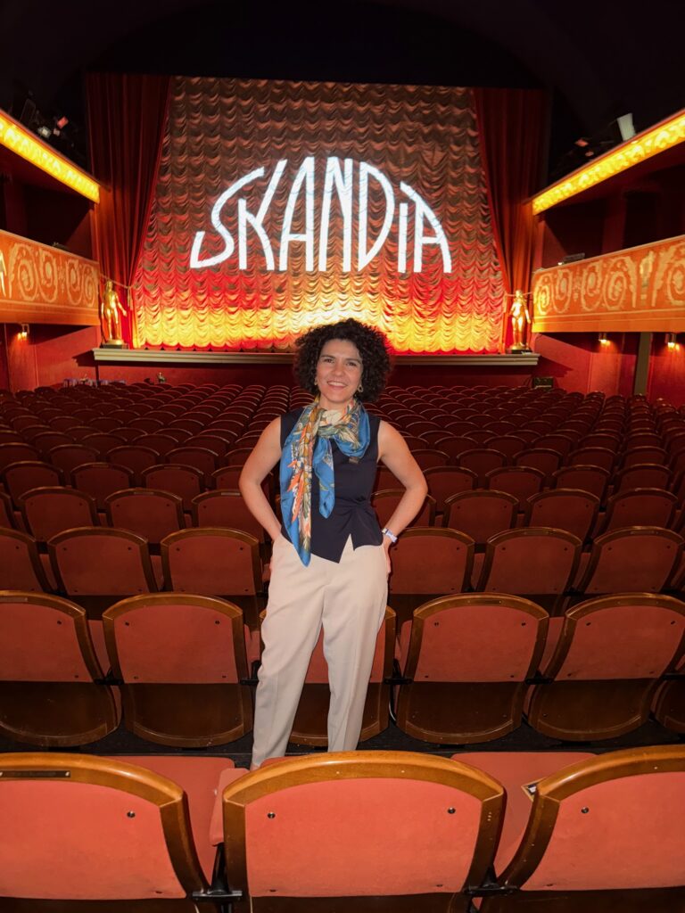 A woman stands smiling in the aisle of an empty theatre with red seats, wearing a colourful scarf and beige trousers. The stage curtain behind her displays the word SKANDIA in large white letters.