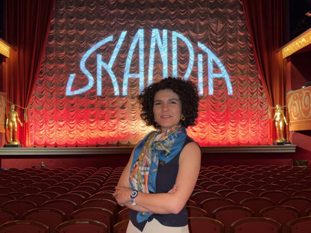 A woman with curly hair stands smiling with arms folded in an empty theatre. The stage curtain behind her is lit up with the word SKANDIA in large letters. The theatre features red seats and ornate decorations.