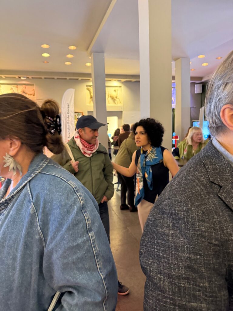 A group of people gather indoors at an event. One woman gestures while talking to a man in a cap and bandana. Others are mingling in the background. The atmosphere appears busy and sociable.