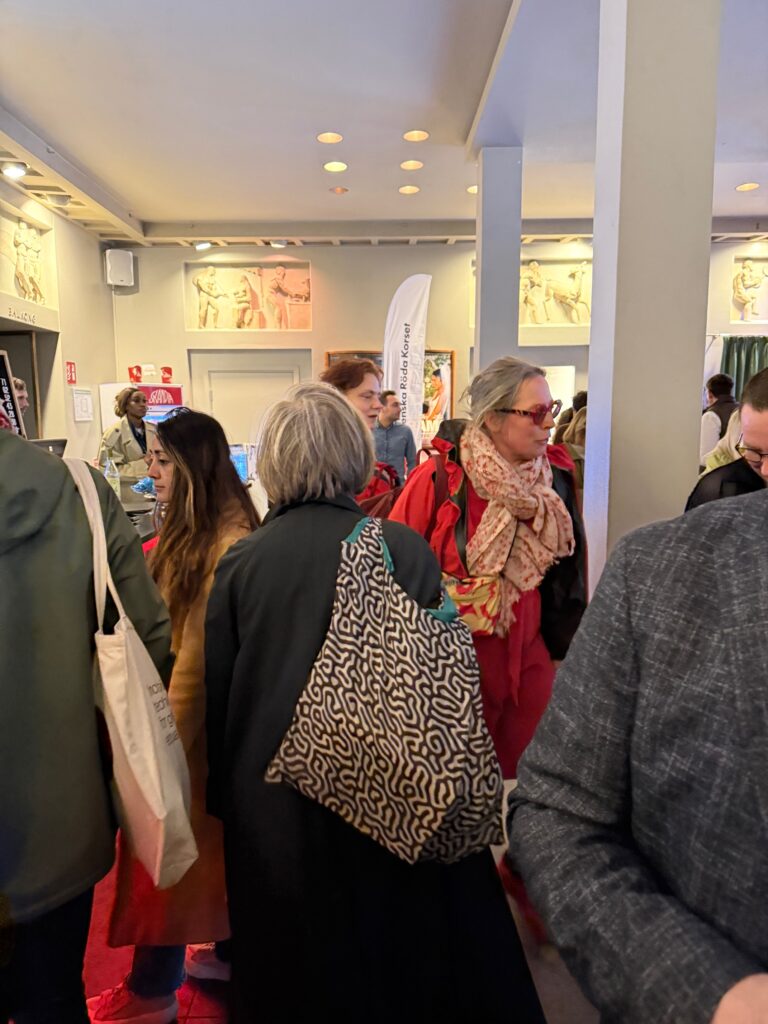 A group of people stand and converse in a brightly lit indoor space with art on the walls; most wear coats and scarves, and one person has a large patterned bag.