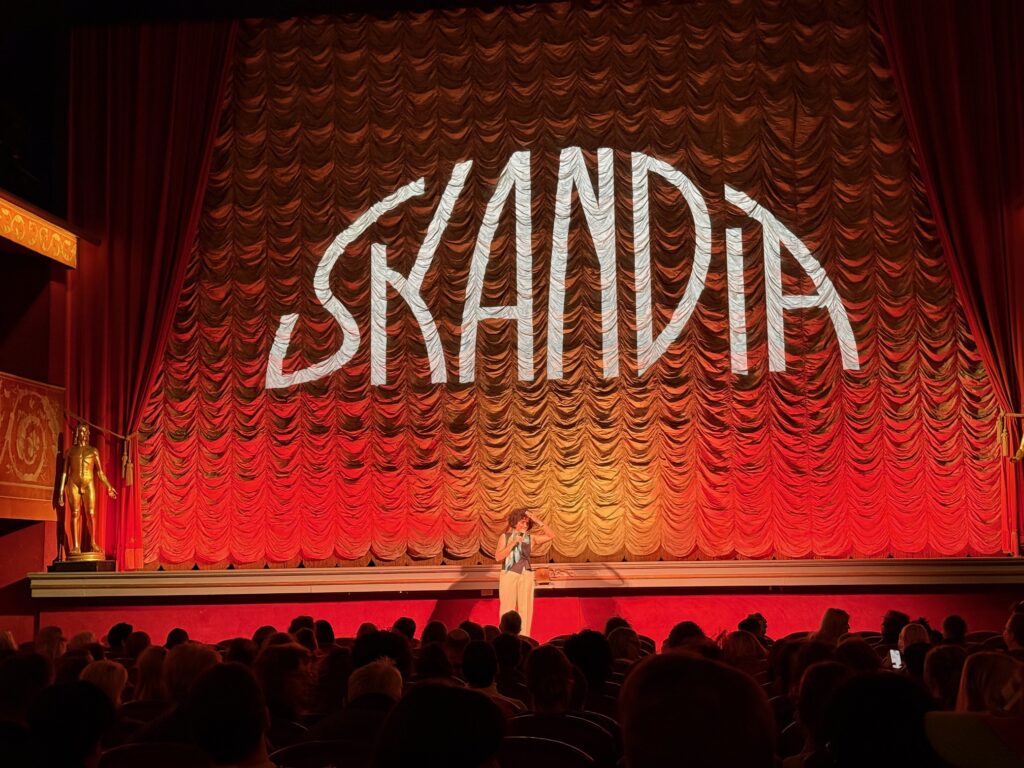 A person stands on stage in front of a large red and gold curtain with the word SKANDIA projected in bold letters, addressing an audience seated in a theatre.