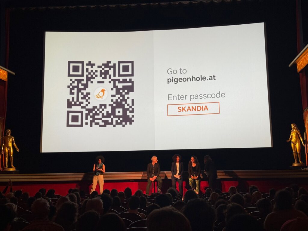 Five people stand on stage in front of a large cinema screen displaying a QR code, instructions to visit pigeonhole.at, and a passcode SKANDIA. The audience is seated, facing the stage.