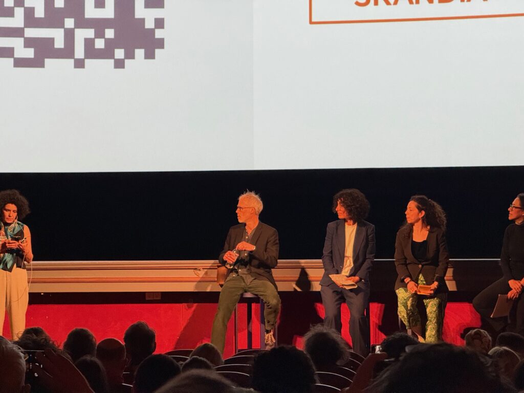 Five people are seated on stage in front of an audience, participating in a panel discussion. A large screen above them displays part of a QR code and some text. The room is dimly lit, with the audience in shadow.