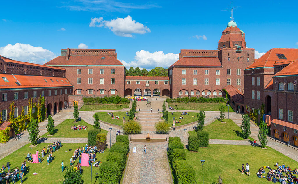 A large red-brick university building with a central tower and arched entrance overlooks a spacious, green courtyard where people are gathered in groups on the grass under a sunny blue sky.