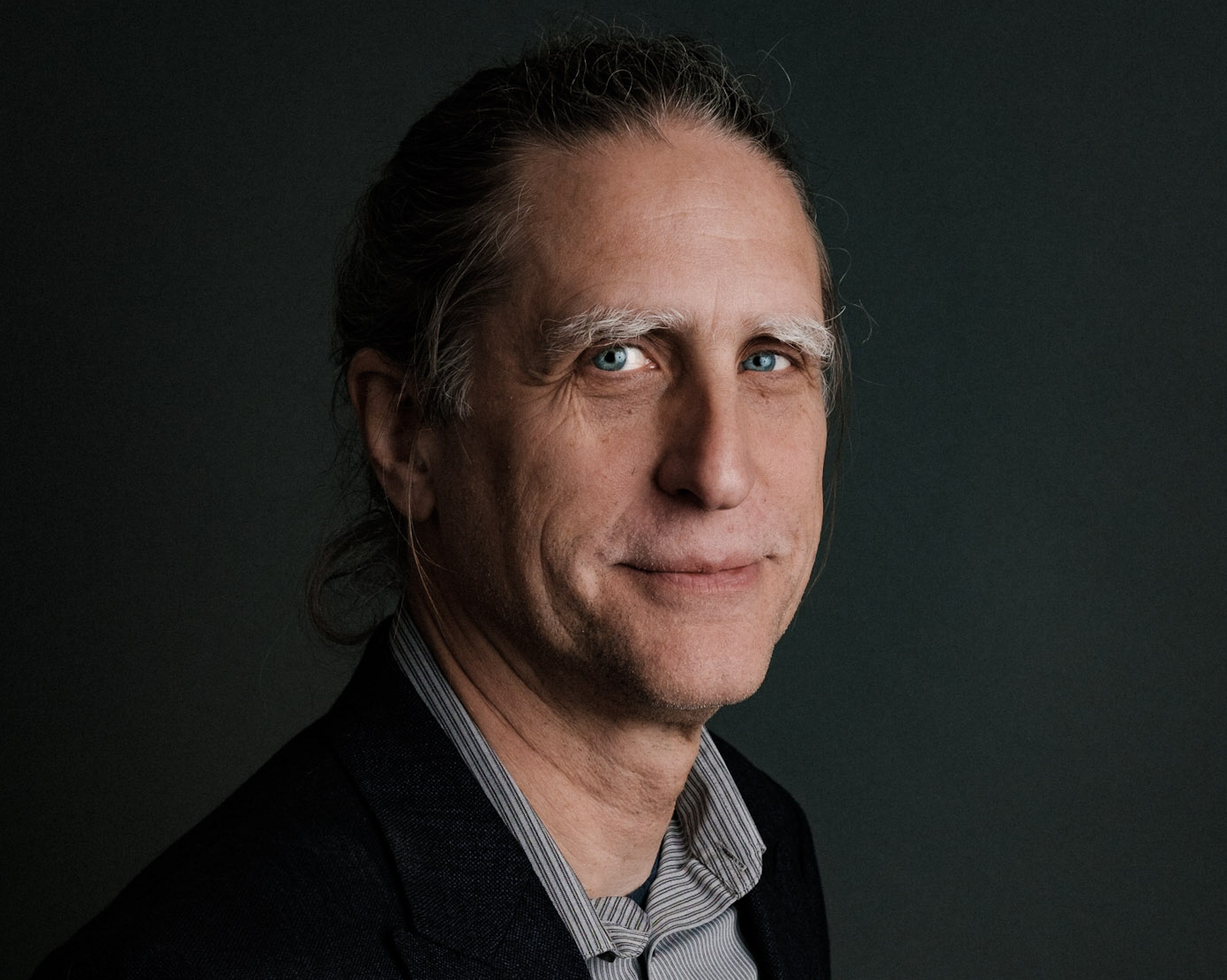 A middle-aged man with long greying hair pulled back, wearing a dark jacket over a striped shirt, smiles slightly against a dark background.