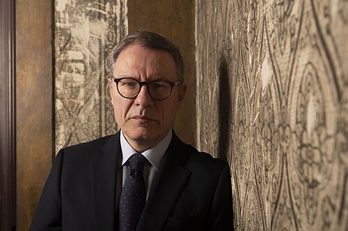 A man in a suit and tie with glasses stands indoors, leaning against a patterned wall. The background features a wooden panel and textured wallpaper. The lighting is dim and the expression is serious.