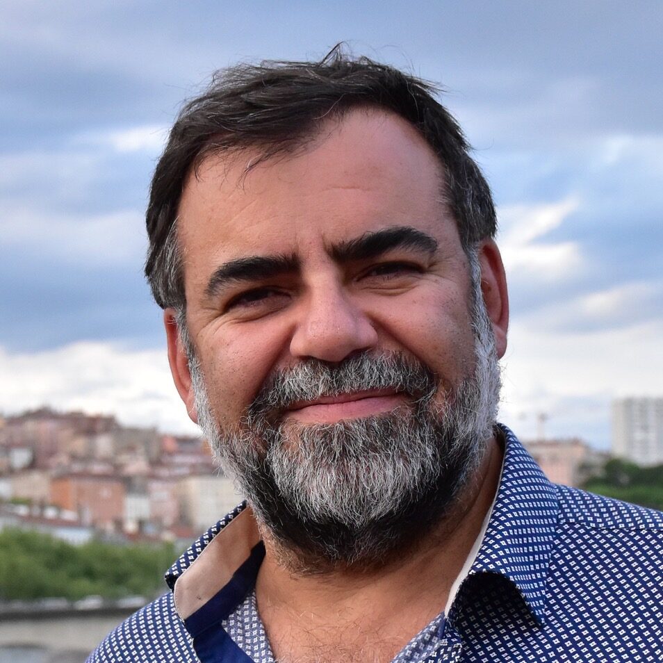 A middle-aged man with a beard and dark hair smiles outdoors, wearing a blue patterned shirt. An urban landscape with buildings and greenery is blurred in the background under a partly cloudy sky.