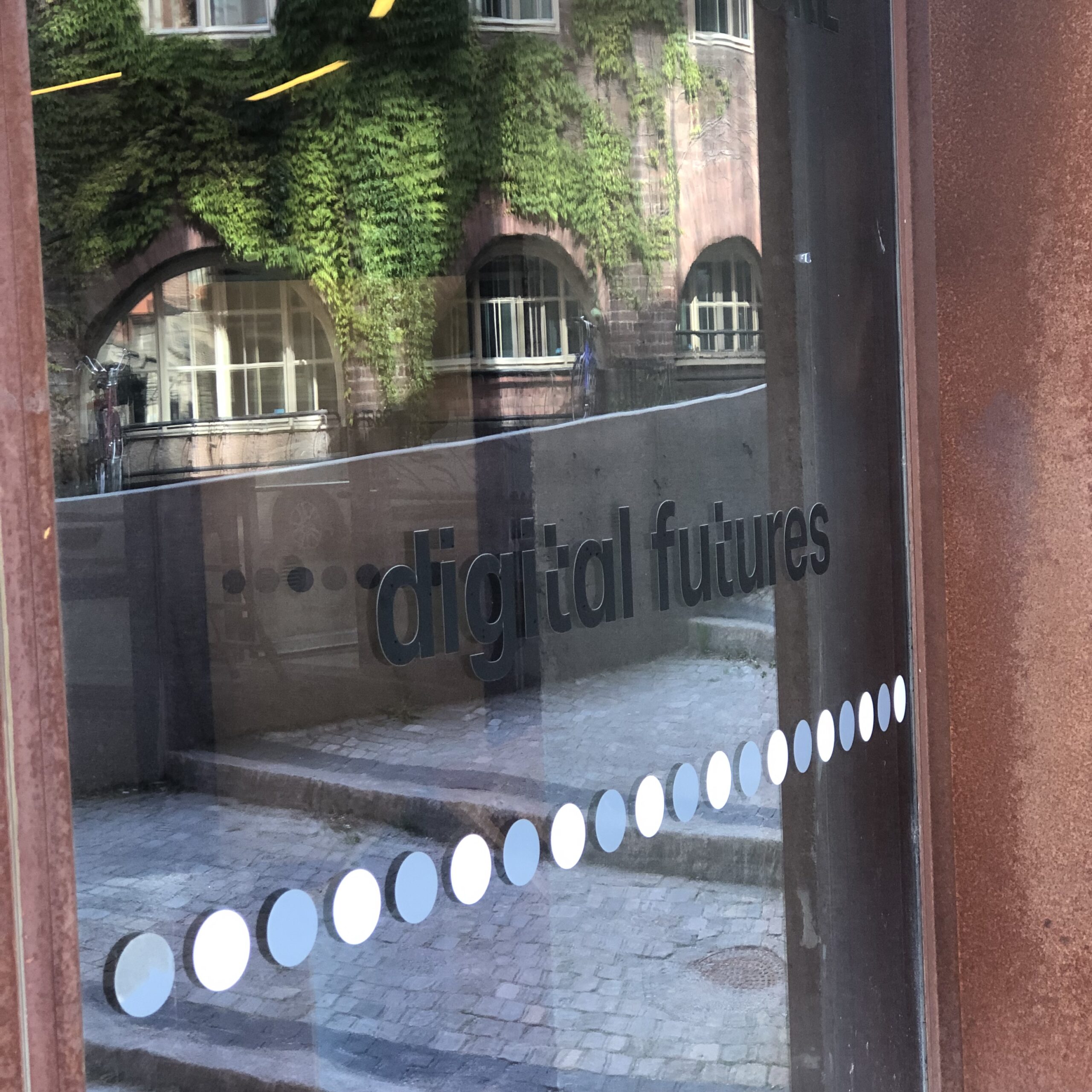 A glass door with the words digital futures and a row of white dots, reflecting an old brick building with green ivy and arched windows across a stone courtyard.