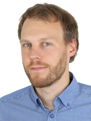 A man with light brown hair, a short beard, and blue eyes is wearing a blue striped button-up shirt and looking slightly to the side. The background is plain white.