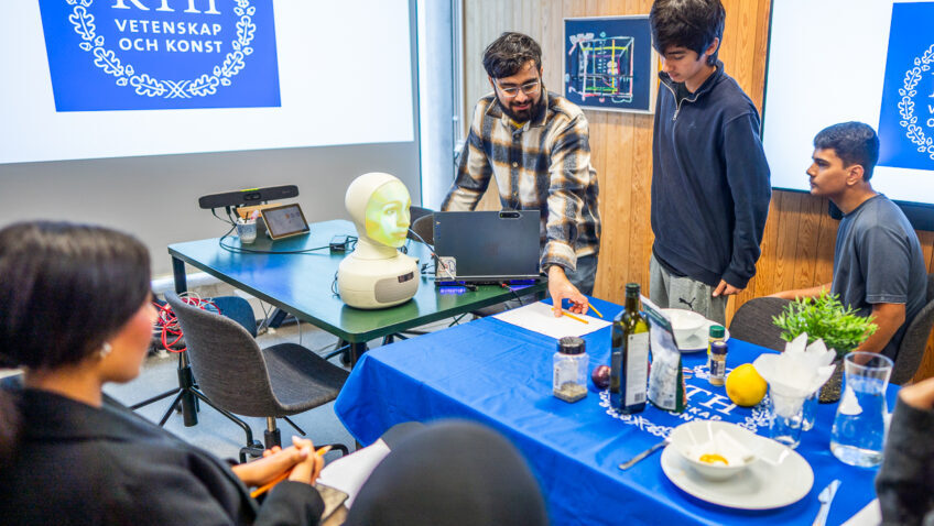 Five students gather around a table set with crockery and props, presenting a project in front of KTH Royal Institute of Technology logos on screens in a wood-panelled room.