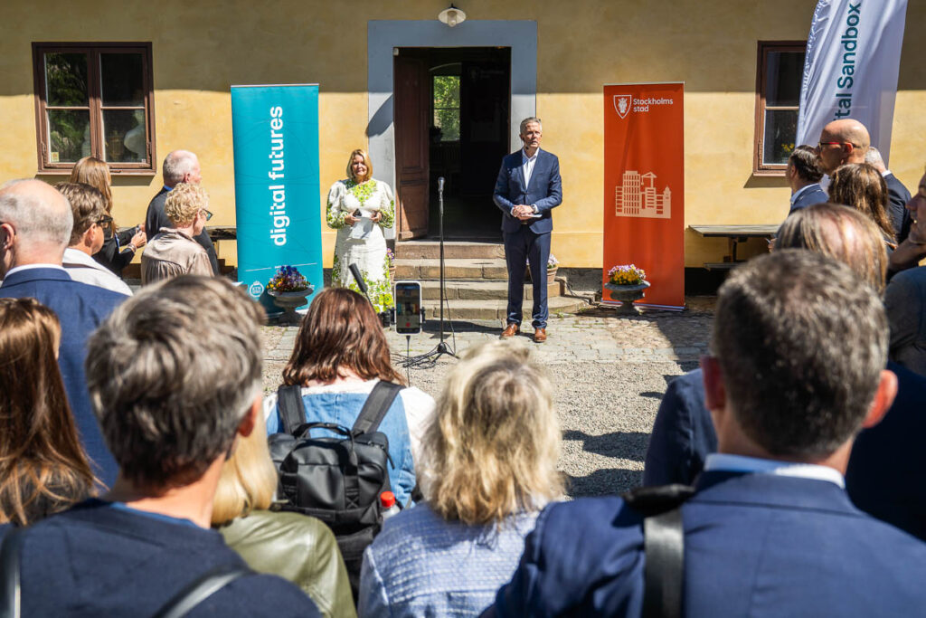A woman and a man speak at microphones in front of an audience outdoors, flanked by colourful banners reading digital futures and Stockholm. The event takes place in front of a yellow building on a sunny day.