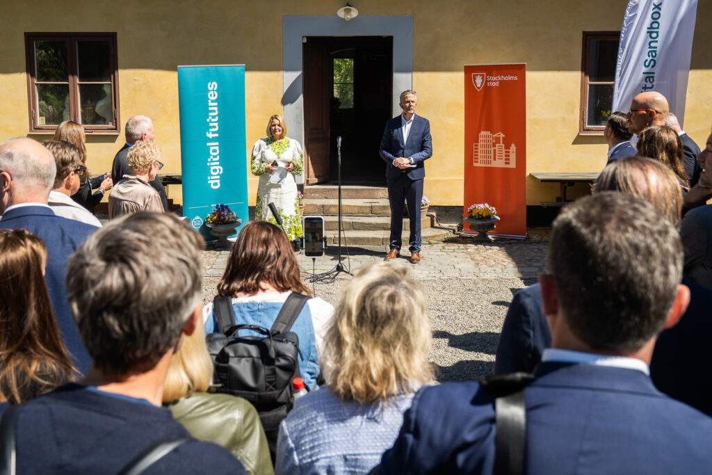 A woman and a man stand on steps, speaking to a crowd outside a yellow building. Banners read “digital futures” and “KTH,” indicating a formal event or presentation. The audience listens attentively in sunny weather.