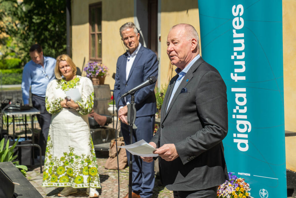 A man speaks into a microphone outdoors near a “digital futures” banner, holding papers. Two people, one in a white dress with green patterns and one in a suit, stand listening in the background.