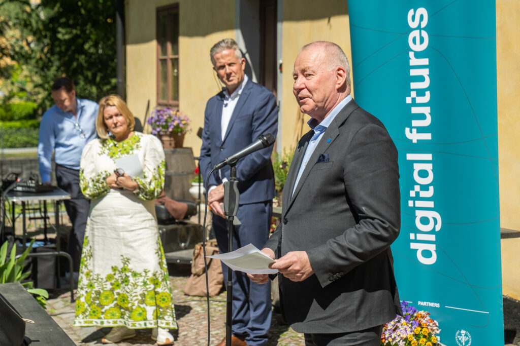 A man in a suit speaks at a microphone outdoors, holding papers. Two people stand behind him, one in a floral dress, another in a suit. A teal banner with digital futures is visible. Flowers and a building are in the background.
