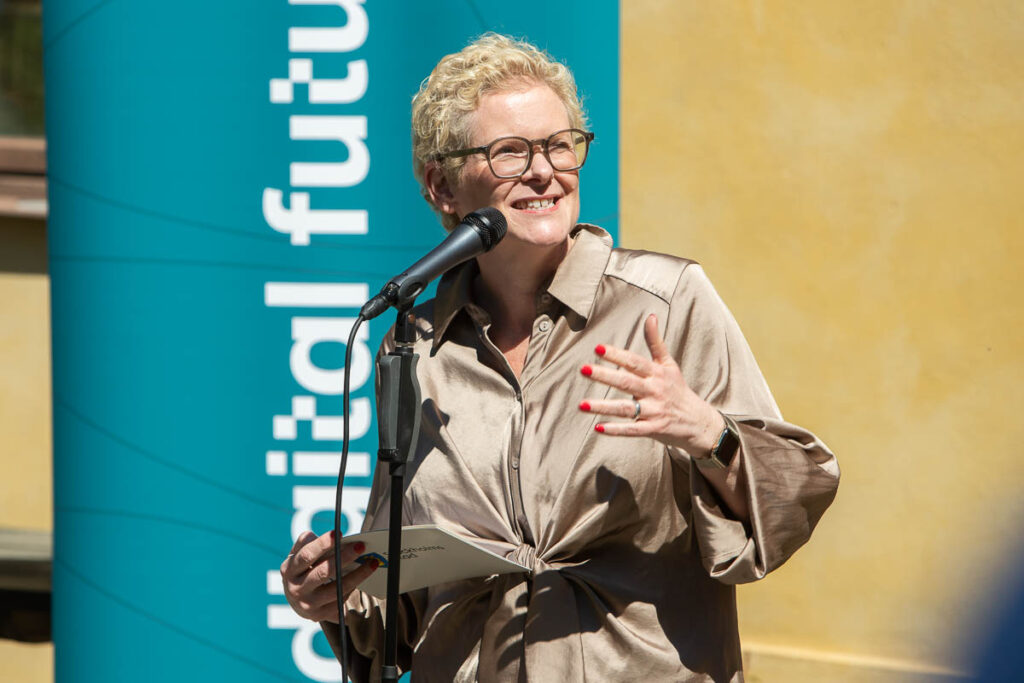 A person with short curly blonde hair and glasses speaks into a microphone outdoors, holding papers. They wear a tan blouse and stand in front of a teal sign that says digital future.