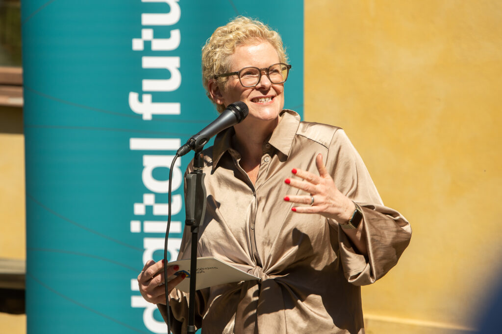 A woman with short blonde hair and glasses speaks into a microphone outdoors, holding papers. She wears a beige blouse and has red nail varnish. A teal banner with partially visible text digital futu is in the background.