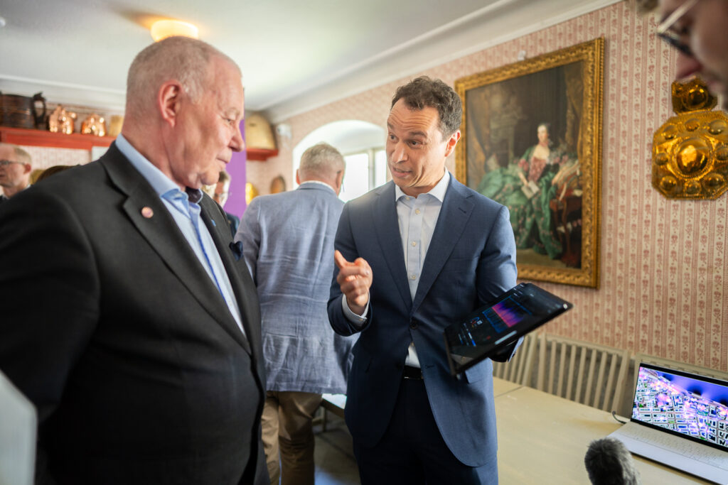 Two men in suits talk indoors; one gestures while holding a tablet. Others are in the background, and a painting and gold decoration are on the wall. A laptop and microphone are visible on a table nearby.