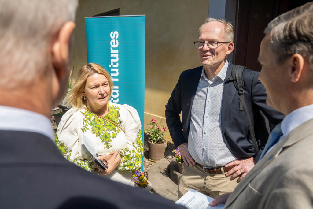 Four people stand outside talking; one woman holds a notebook and faces three men in suits. A turquoise banner with partially visible text is in the background, along with a plant and a yellow wall.