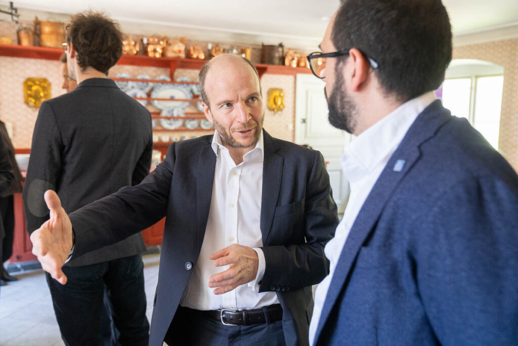 A man in a suit gestures with his hand while speaking to another man in glasses and a blazer in a well-lit room with shelves and copper pans in the background.