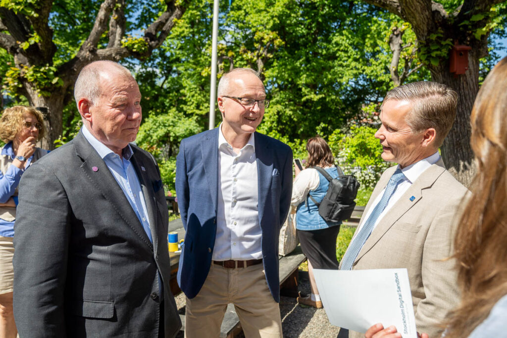Three men in suits are talking and smiling outdoors on a sunny day, with trees and other people in the background. One man holds a document. The mood appears friendly and informal.
