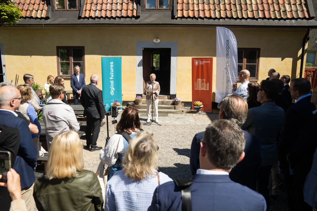 A woman speaks at a lectern outside a yellow building, surrounded by an audience. Two banners reading digital futures and Stockholm University stand beside her. The event takes place on a sunny day.