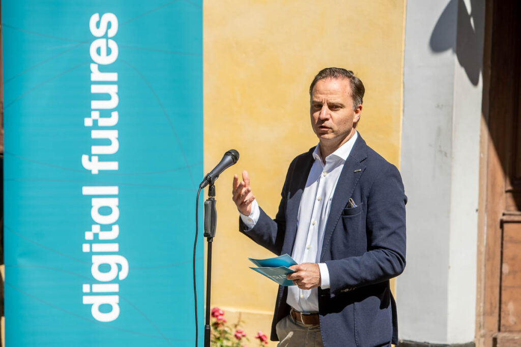 A man in a navy blazer speaks at an outdoor event, standing beside a vertical banner that reads digital futures. He gestures with one hand and holds papers in the other, with a microphone in front of him.