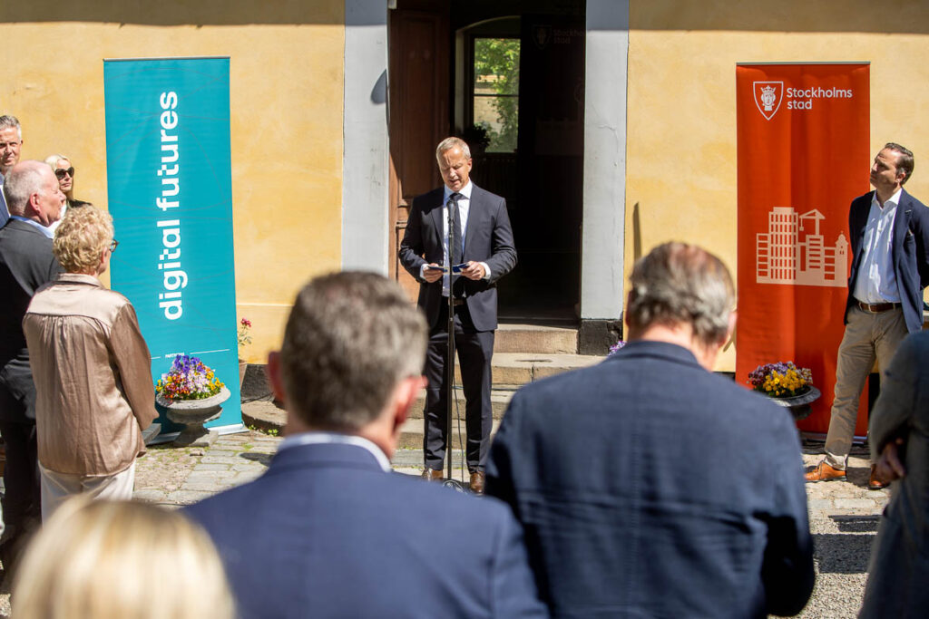 A man speaks at a microphone outdoors to a small audience, flanked by blue and orange banners reading “digital futures” and “Stockholms stad.” The event takes place in front of a yellow building on a sunny day.