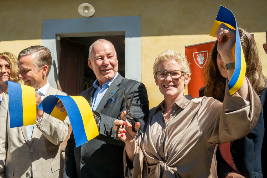 A group of smiling people stand together at a ribbon-cutting ceremony, holding up blue and yellow ribbons. One person holds large scissors, about to cut the ribbon. The scene is outside, in front of a doorway.