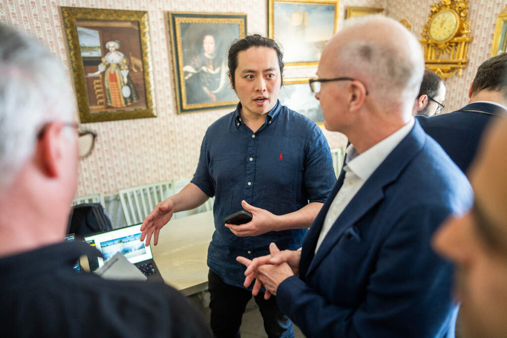 A man in a blue shirt is speaking and gesturing with his hands to a group of people in a room decorated with framed paintings and an ornate clock.