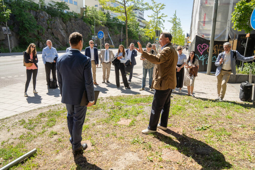 A group of people in business attire stand outdoors on a sunny day, gathered around a speaker who gestures while talking. They are on a pavement near a street, with trees and buildings in the background.