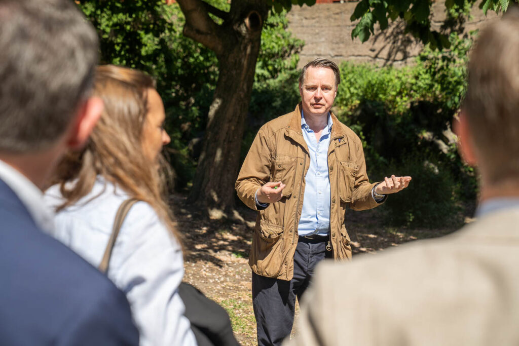 A man in a brown jacket gestures whilst speaking outdoors to a small group of people, with trees and greenery in the background on a sunny day.