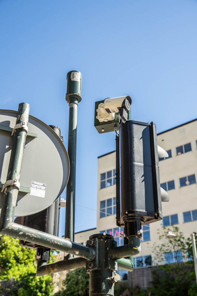 A close-up view of a traffic camera mounted on a pole next to a traffic light, with a building and clear blue sky in the background.