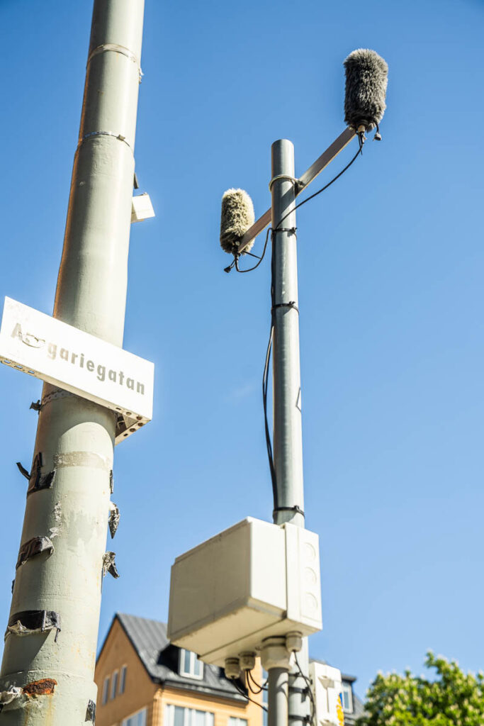 Two microphones with windshields mounted on tall poles next to a street sign reading Åsögatan, with a blue sky and part of a building visible in the background.