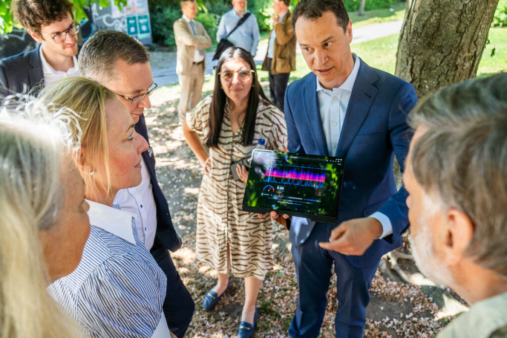 A group of people stand outdoors as a man in a suit shows them a tablet displaying colourful graphs. The group appears engaged, with trees and grass in the background.