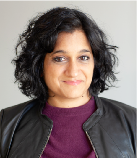 A person with wavy, dark hair wearing a black jacket over a maroon top, smiling softly whilst looking at the camera against a plain light background.