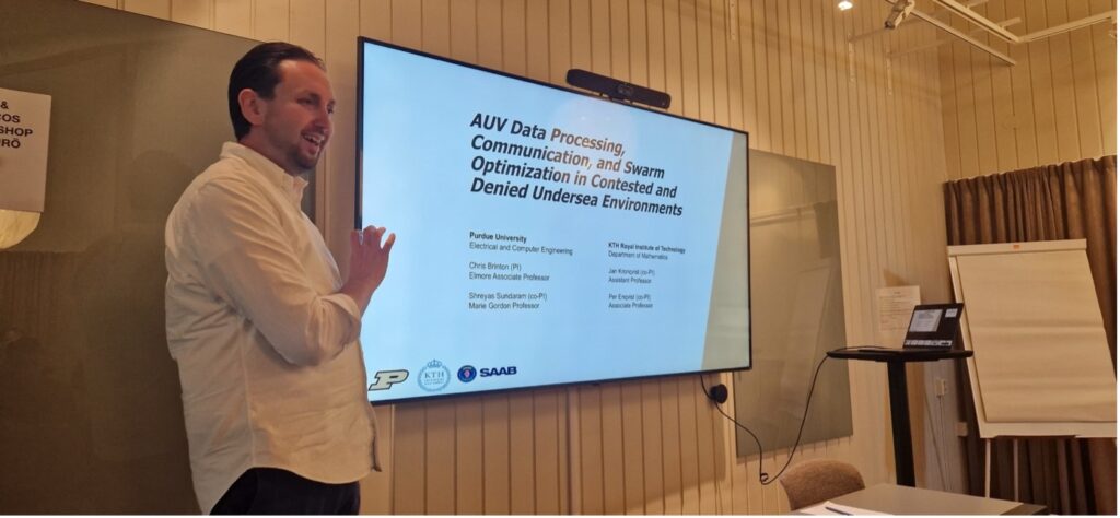 A man stands and gestures towards a large screen displaying a presentation titled AUV Data Processing, Communication, and Swarm Optimisation in Contested and Denied Undersea Environments in a meeting room.