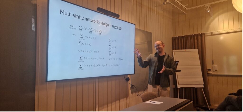 A man is giving a presentation in a conference room, pointing at a large screen displaying mathematical equations under the title Multi-static network design (ongoing). A flip chart is in the background.