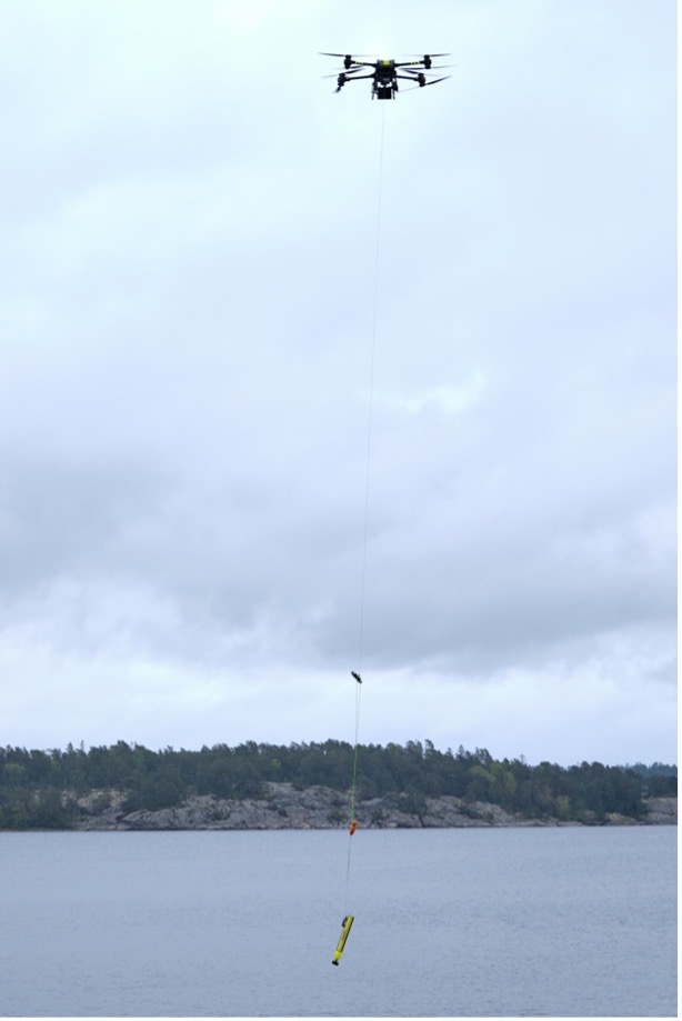 A drone hovers over a body of water, lowering a cylindrical object on a long line, with a shoreline and trees visible in the background under a cloudy sky.