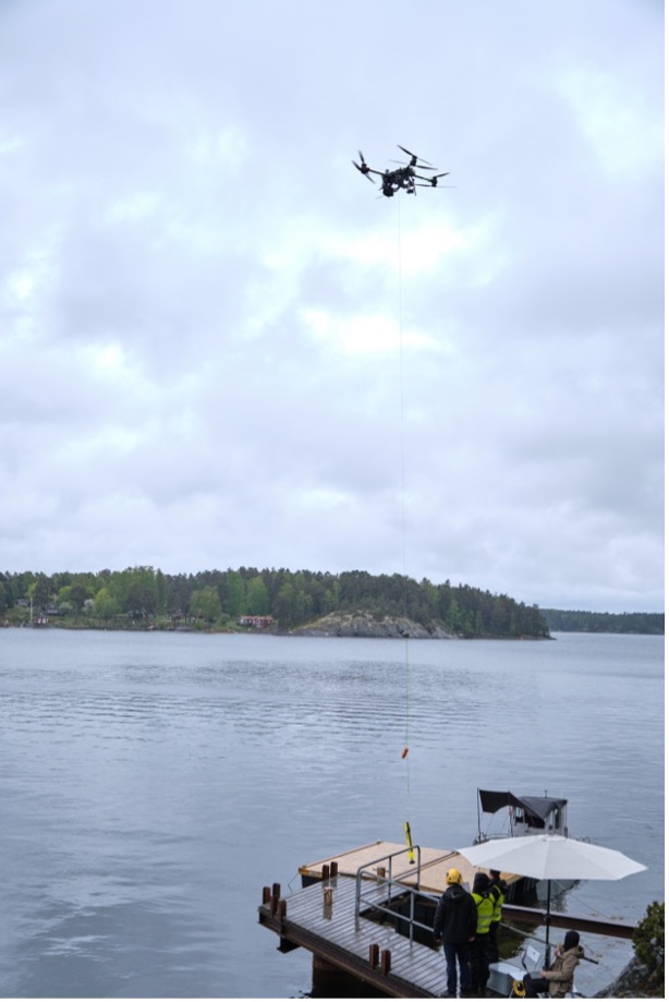 A drone hovers above a lakeside jetty, lowering an object on a cord while two people in yellow safety vests and helmets stand on the jetty under an umbrella. The lake and trees are visible in the background.