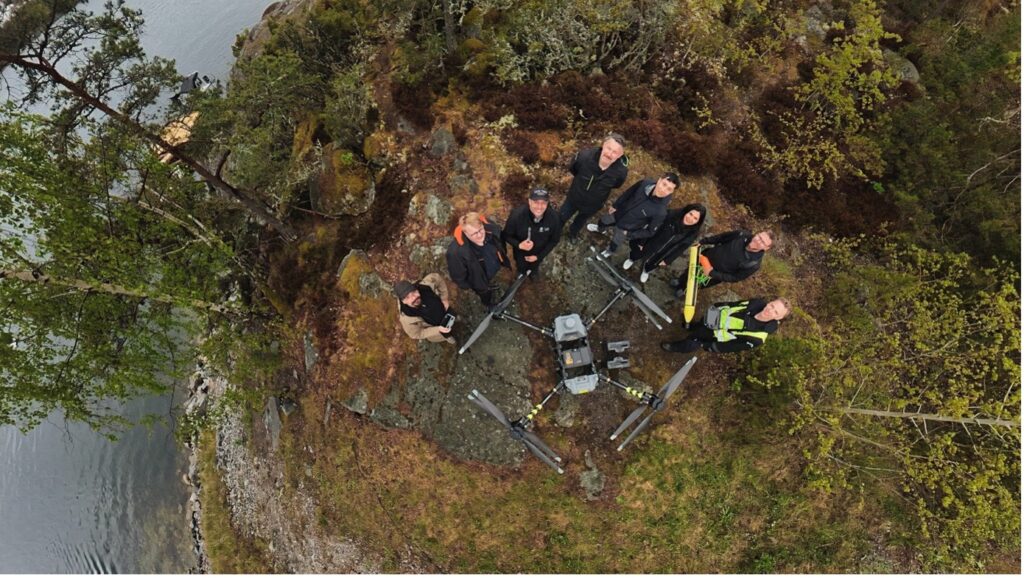 Aerial view of eight people standing on a rocky, forested cliff near water, looking up at the camera. Several drone devices and equipment are arranged on the ground in front of the group.