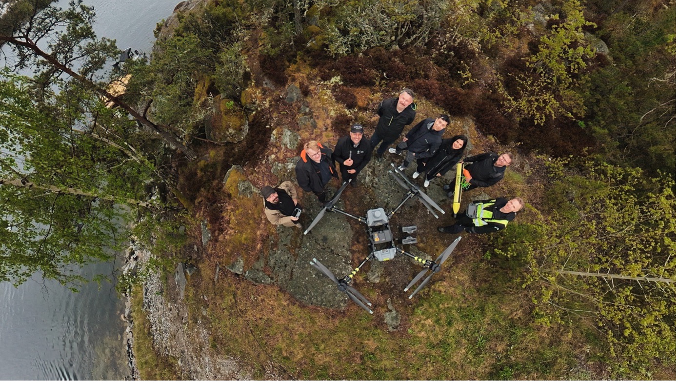 Aerial view of eight people standing on a rocky, forested cliff near water, looking up at the camera. Several drone devices and equipment are arranged on the ground in front of the group.
