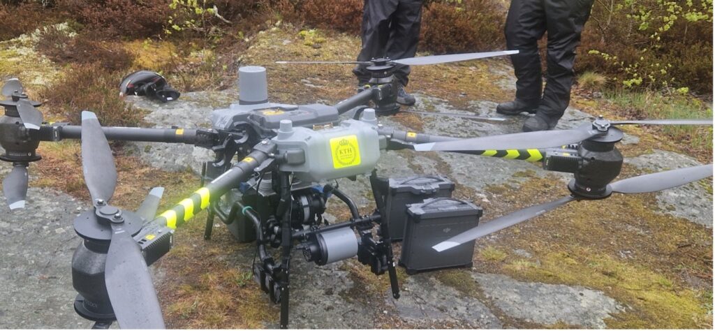 A large industrial drone with four rotors, cameras, and sensors is on the ground outdoors. Two people in black clothing stand nearby, and foliage is visible in the background.