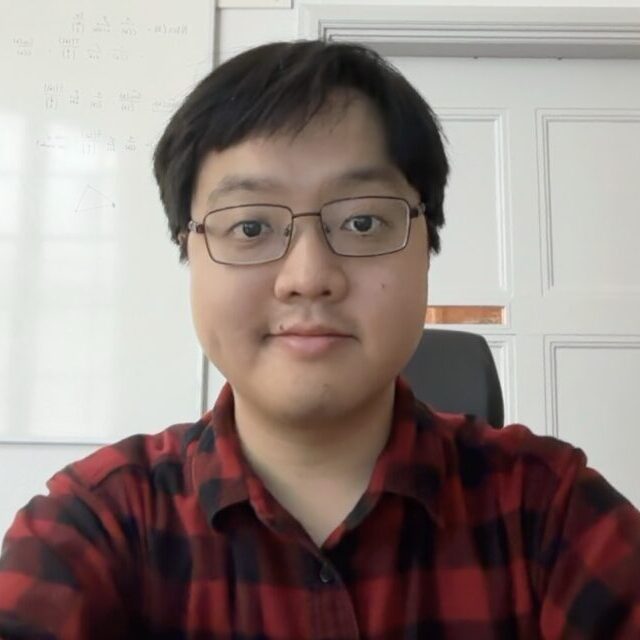 A person wearing glasses and a red checked shirt sits indoors, smiling at the camera. A whiteboard with diagrams and equations is visible on the wall behind them.
